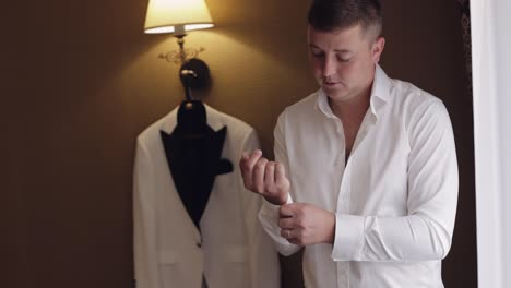 handsome groom man dresses in wedding morning and fixes his buttons on a white shirt sleeve, indoors