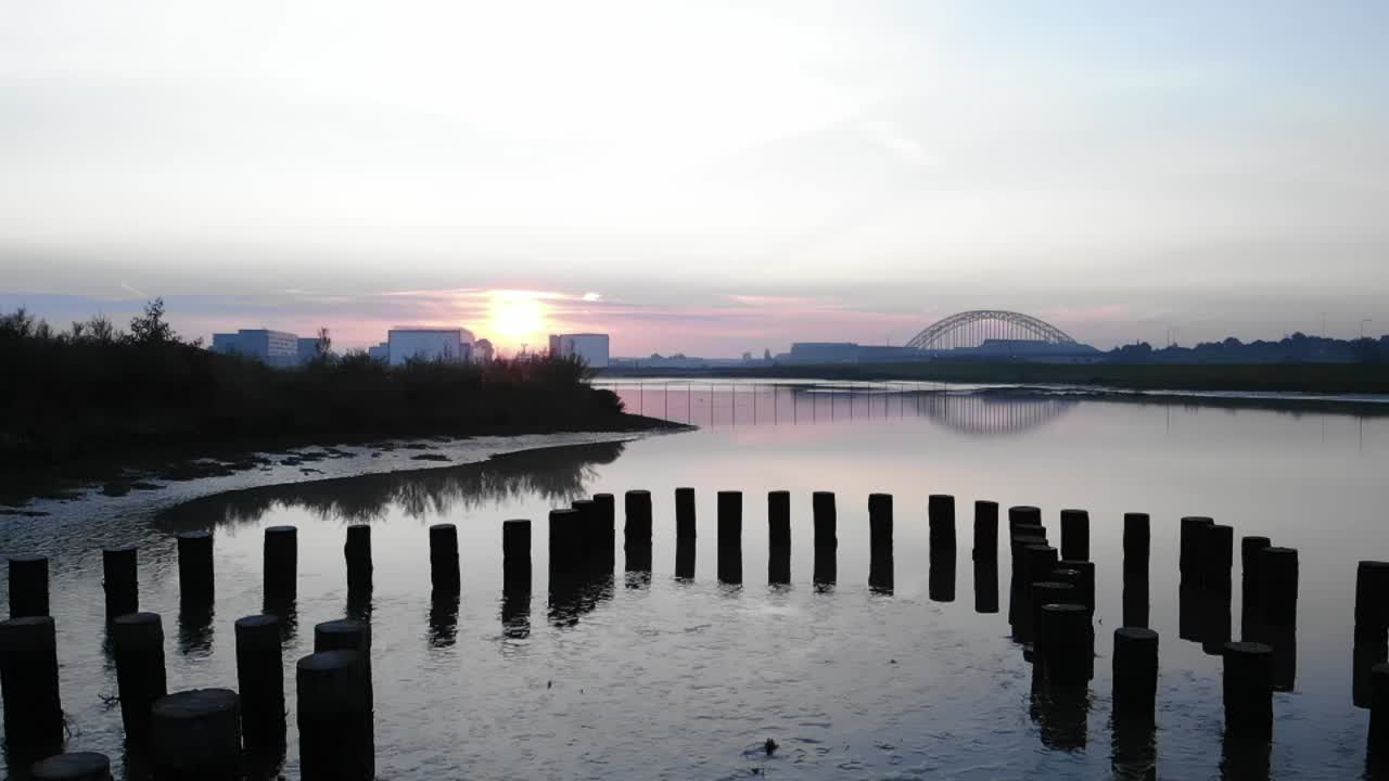 강둑에 있는 물과 나무에 실루엣이 있는 나무 기둥, 일몰에 아치교가 있는 멋진 도시, 드론 공중 높은 프레임 속도 - 느린 모션