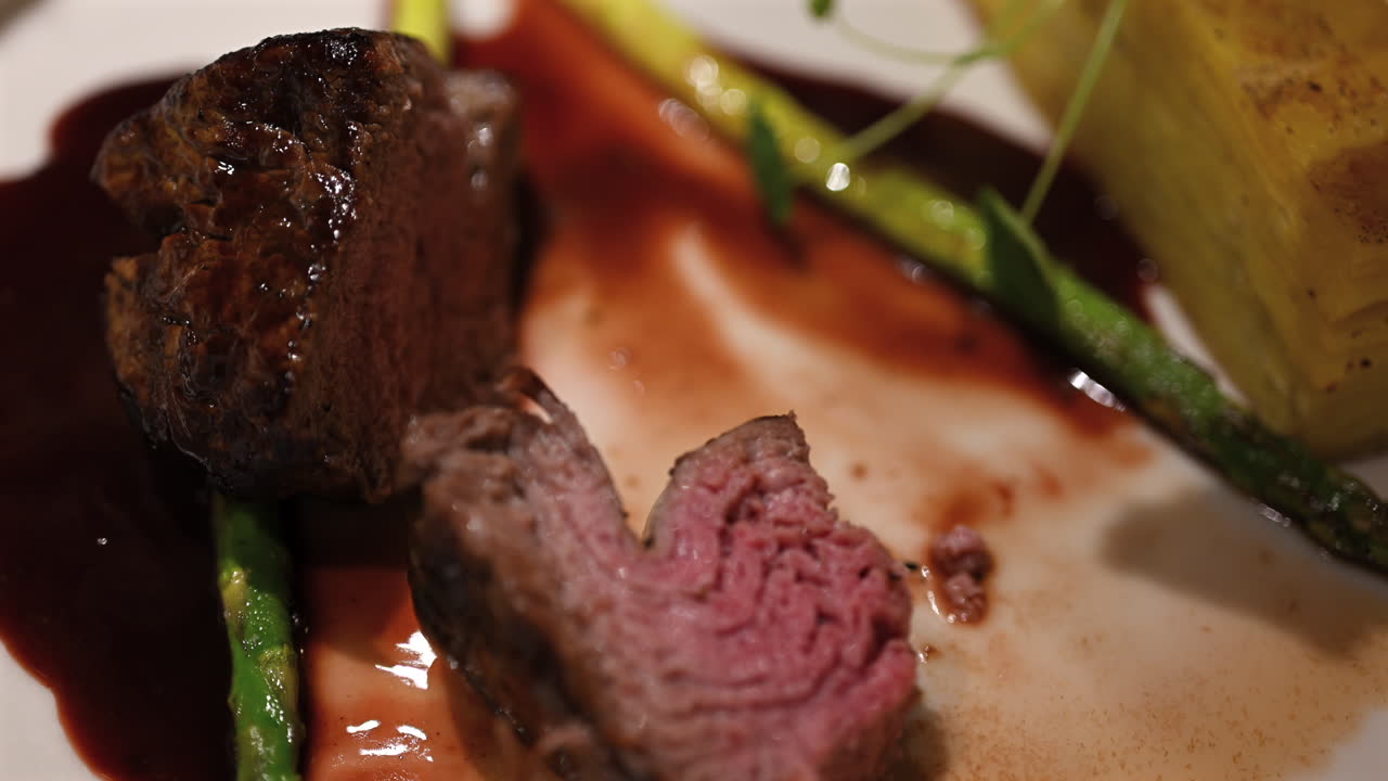 Primo piano di una donna che taglia una succulenta bistecca di manzo gourmet con coltello e forchetta su un elegante piatto da ristorante con asparagi e salsa.