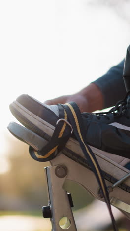 primer plano de la mano masculina ajustando las correas de los zancos con zapatillas de deporte negras y azules visibles. lleva una sudadera gris con capucha, con la luz del sol iluminando suavemente la escena y la vegetación borrosa en el fondo
