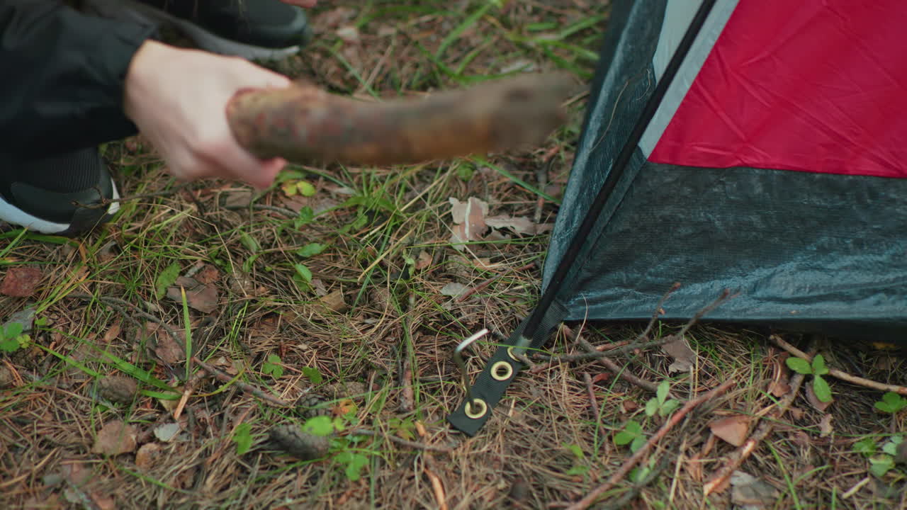 partial view of girl securing tent base by driving metal peg through grommet using thick wooden stick while squatting on forest ground with scattered twigs, dry leaves, and pine needles around