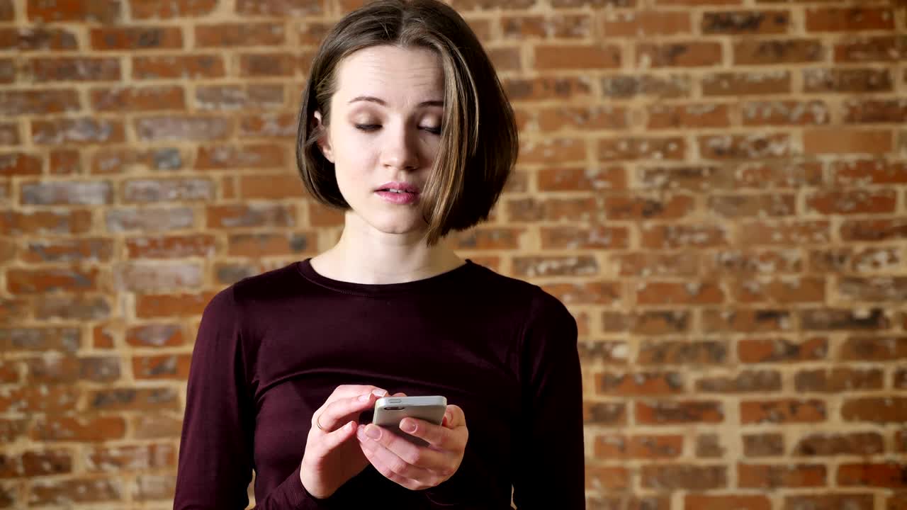 una joven alegre está escribiendo un mensaje en su teléfono inteligente, mirando a la cámara, sonriendo, concepto de comunicación, fondo de ladrillo