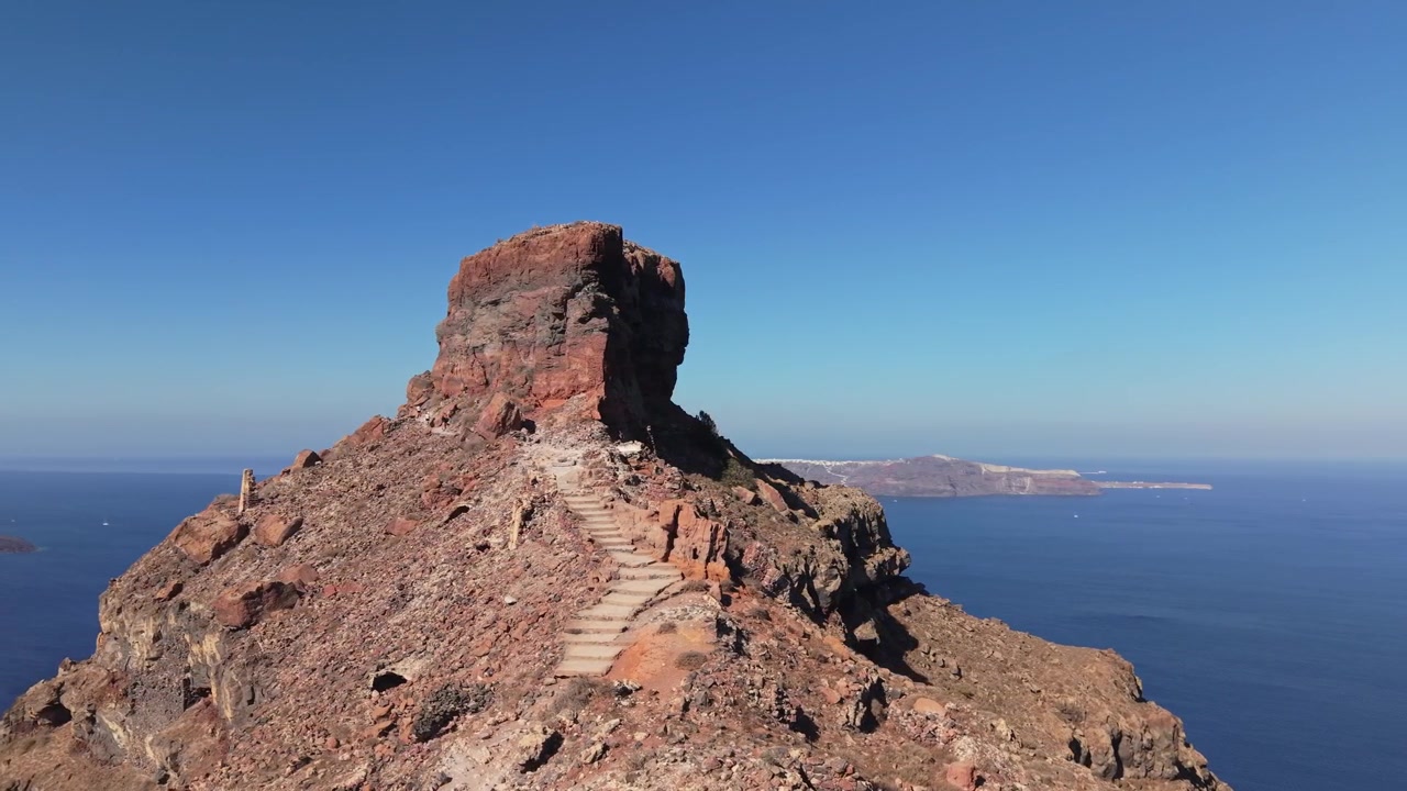 La Roccia di Skaros in primo piano con il paesaggio vulcanico di Santorini sullo sfondo, che mostra le scogliere drammatiche della caldera e il profondo blu del Mar Egeo.
