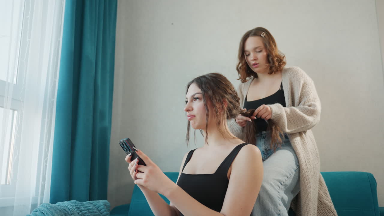 Relaxed Livingroom White Women Styling Hair, Teal Curtain Backdrop, Natural Window Light, Seated Client Checking Phone, Standing Friend Finishing Braid, Informal AtHome Salon Vibe, Modern Lifestyle