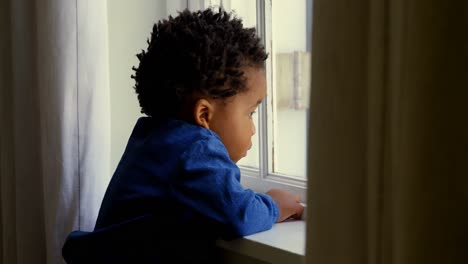 Vista-Lateral-De-Un-Lindo-Niño-Negro-Apoyado-En-El-Alféizar-De-La-Ventana-En-Una-Cómoda-Casa-4k