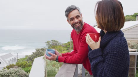 feliz pareja diversa bebiendo café y hablando juntos en el balcón