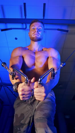 healthy athlete pumping up his arm muscles. sportsman without shirt is training biceps on simulator on blue gym background. view from below. vertical video