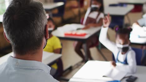 maestro masculino diverso en el aula con niños levantando las manos durante la lección, todos con máscaras faciales