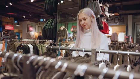 curious window shopper with platinum hair in chunky white turtleneck sweater glides fingers across shearling coat rack, comparing textures while another customer shops in brightly lit fashion store