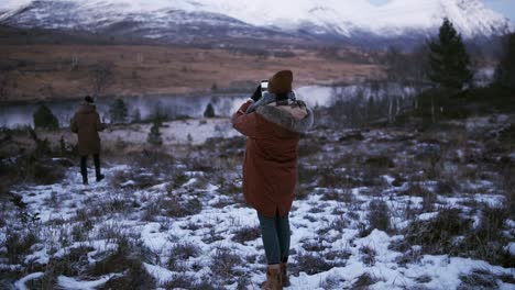 dos turistas caminando en cámara lenta por el campo hacia los picos nevados del fondo. una persona con un abrigo de invierno tratando de tomar una foto de la hermosa naturaleza noruega