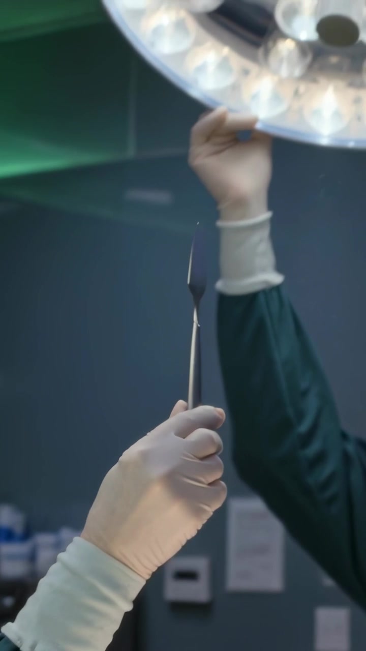 Vertical video: Holding scalpel by surgeon in OR as tech in scrubs adjusting LED lamp for prep