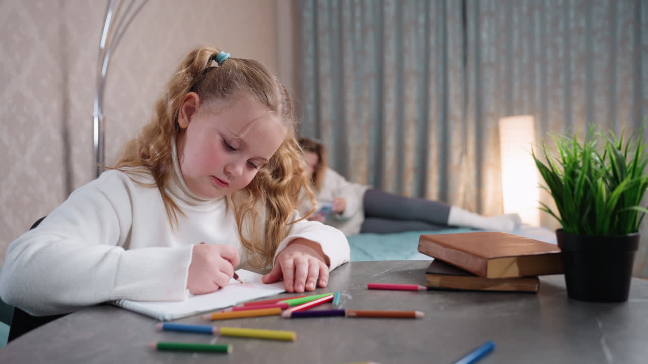 student concentrating while drawing with yellow pencil at table, colorful pencils scattered, books and plant beside her, mother relaxing on bed in background, cozy room setting family lifestyle