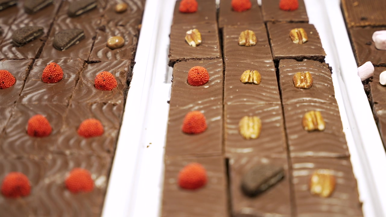 Brownies al cioccolato rettangolari guarniti con caramelle rosse, noci pecan e biscotti, allineati su vassoi bianchi in una vetrina di pasticceria.