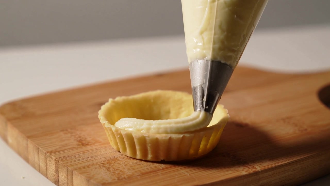 Mano di pasticcere che riempie un guscio di crostata scanalato su tagliere da cucina con beccuccio di metallo per dessert