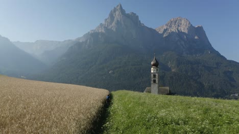 Cyclist-cycling-in-between-two-colored-fields-next-to-a-white-church-and-mountain-background-at-Kastelruth,-North-Italy,-Trentino