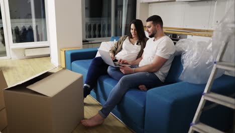 mujer embarazada y un hombre positivo están usando una computadora portátil sentada en el sofá en una nueva sala de estar