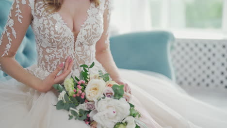 stylish young bride sits on a couch and holds a beautiful tender flowers. young model in white wedding dress is sitting in the light room.