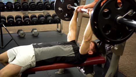 Teen-male-bodybuilder-wearing-black-shirt-lying-down-and-pressing-large-barbell-as-a-man-in-organge-spots-him