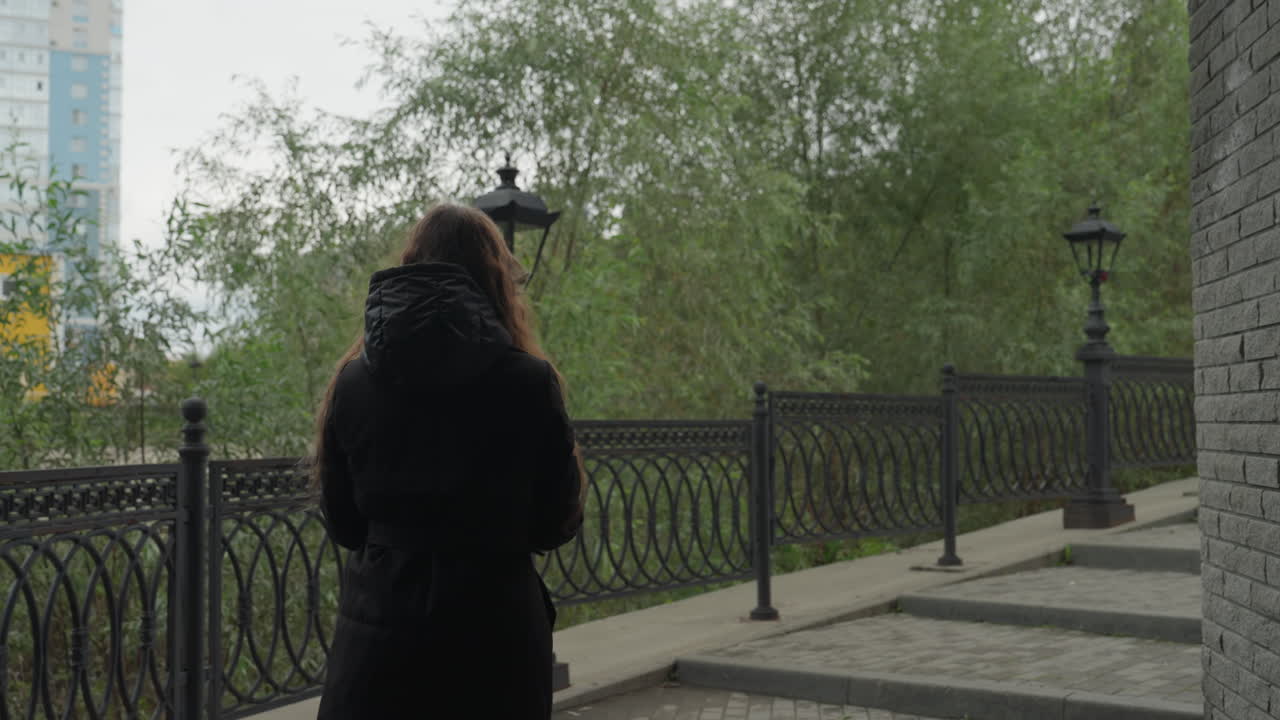 young woman walking with umbrella riverside, overcast sky, iron railing and vintage lampposts, wet pavement and autumn foliage, contemplative mood, turning and closing umbrella while continuing