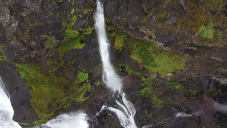 Aerial-tilt-up-shot-of-powerful-waterfalls-between-rocky-and-mossy-mountains-in-Icelandic-Scenery