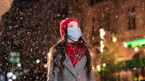 close-up view of cheerful little girl wearing facial mask standing on street in winter and trying to catch snow in christmas