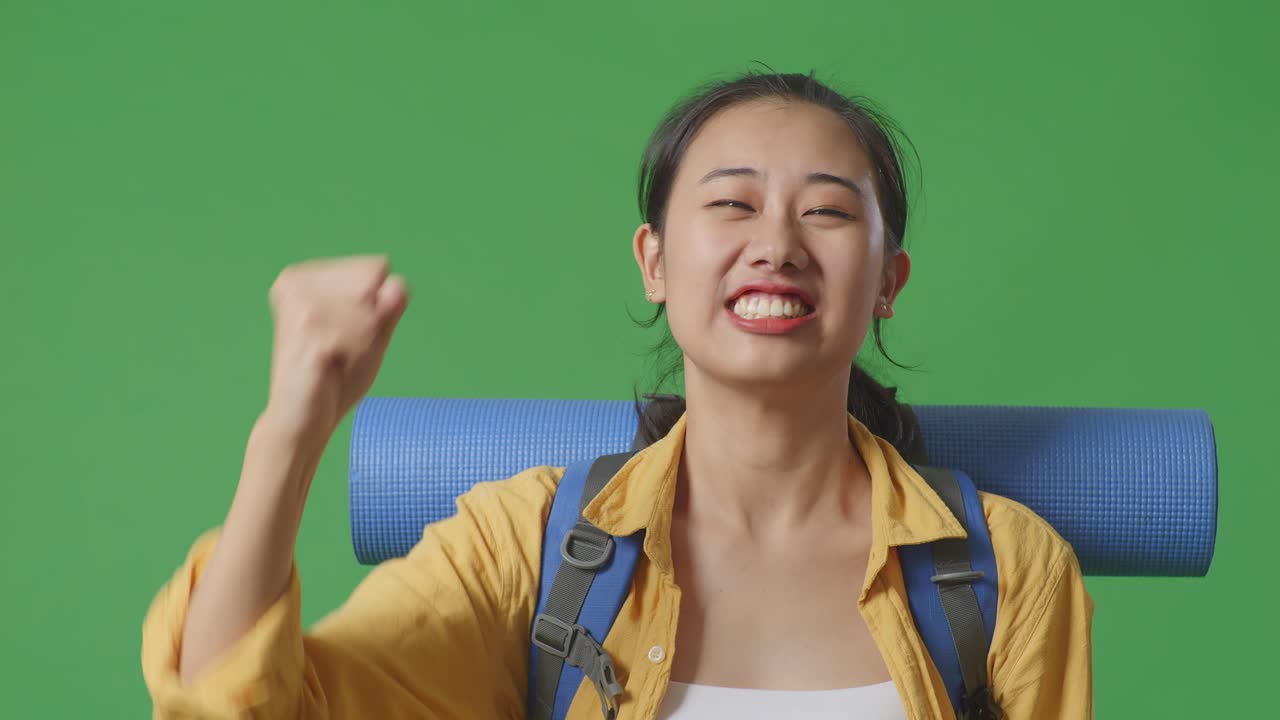 아시아 여성 하이커가 산악 배을 들고 성공을 축하하는 목표를 외치며 스튜디오에서 녹색 화면 배경에 서 있습니다.