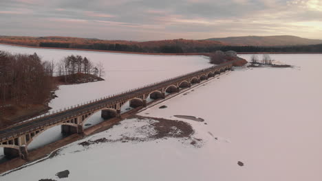 Vuelo-Aéreo-Sobre-El-Puente-Arquitectónico-En-El-Lago-Congelado-En-La-Colorida-Luz-Del-Atardecer