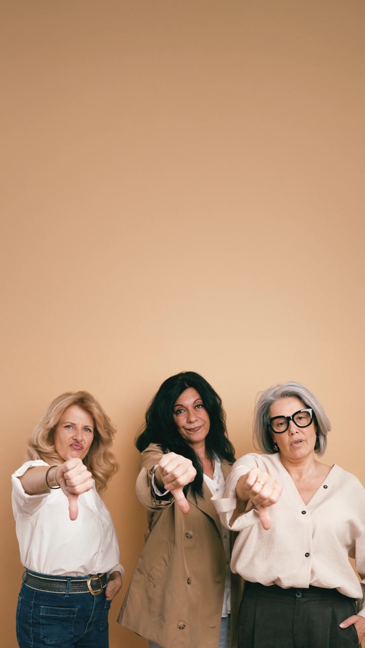Three Mature Women Giving Thumbs Down Gesture Against Beige Background