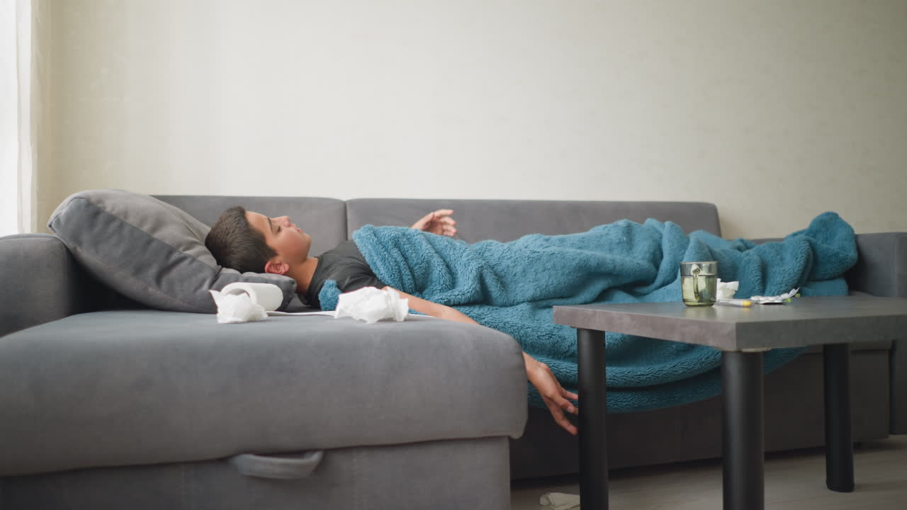 Side view of child laying on couch covered with blanket adjusting for comfort, glass cup on table in living room, peaceful indoor setting with natural light streaming through the window