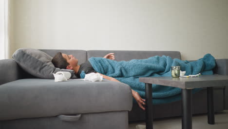 side view of child laying on couch covered with blanket adjusting for comfort, glass cup on table in living room, peaceful indoor setting with natural light streaming through the window