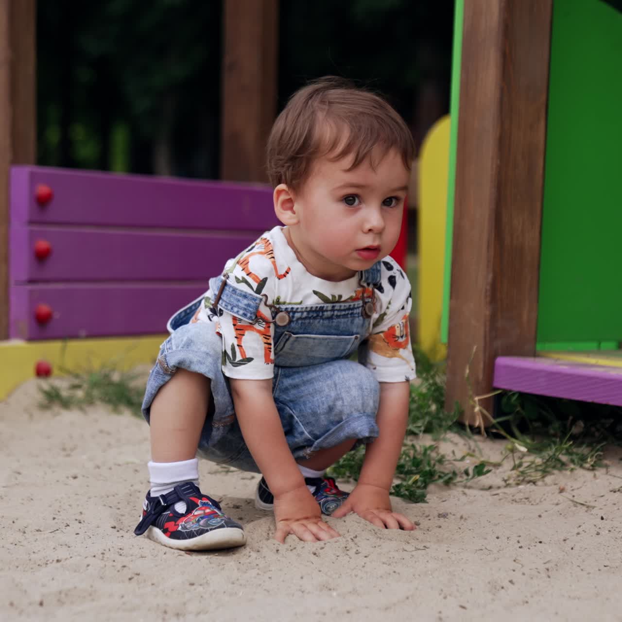 Caucasian kid in romper touching the sand on the playground. Little boy deepens hands into the sand and looking around