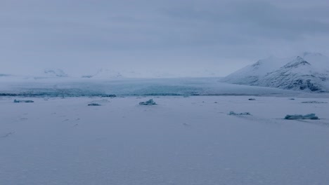 Aerial-panoramic-landscape-view-of-the-frozen-Jokulsarlón-lake-area,-with-icebergs-covered-in-snow,-in-Iceland,-at-dusk