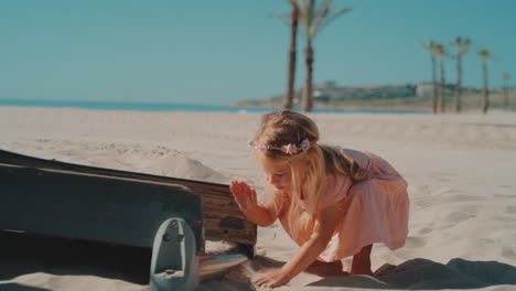 mała dziewczynka o blond włosach i kwiatach we włosach bawiąca się rękami na piasku na plaży w słoneczny dzień