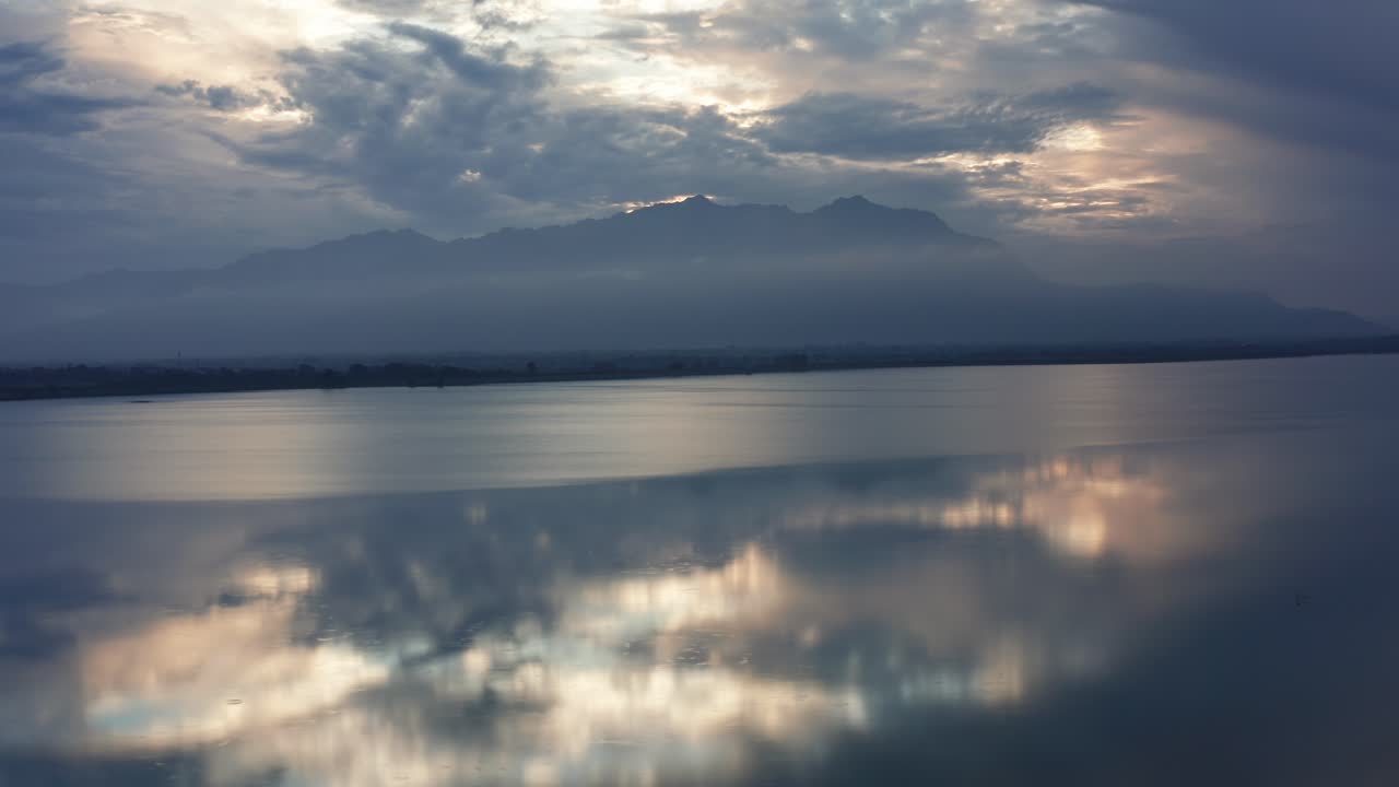 Slow sideways drone movement reveals serene Banas Dam waters reflecting dramatic sunset clouds and distant hazy Mount Abu mountains in Rajasthan's Aravalli range