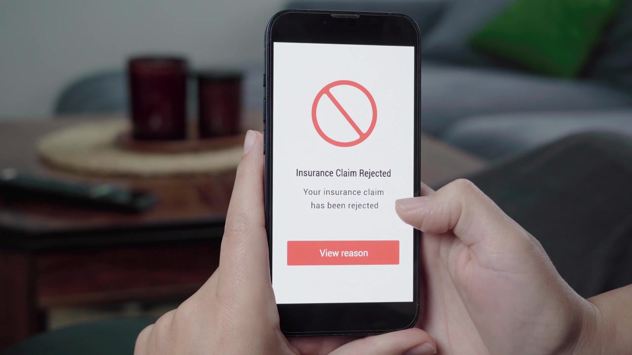 Hands hold a phone as an Insurance Claim Rejected message appears with a red prohibition icon and View reason button.