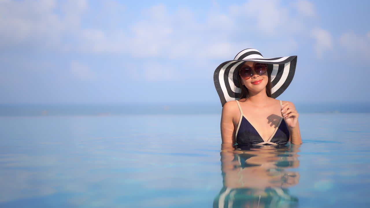 Woman in black bathing suit, sunglasses and large hat standing in water of infinity swimming pool at luxury tropical resort with ocean in background. Attractive Asian tourist enjoying island holiday