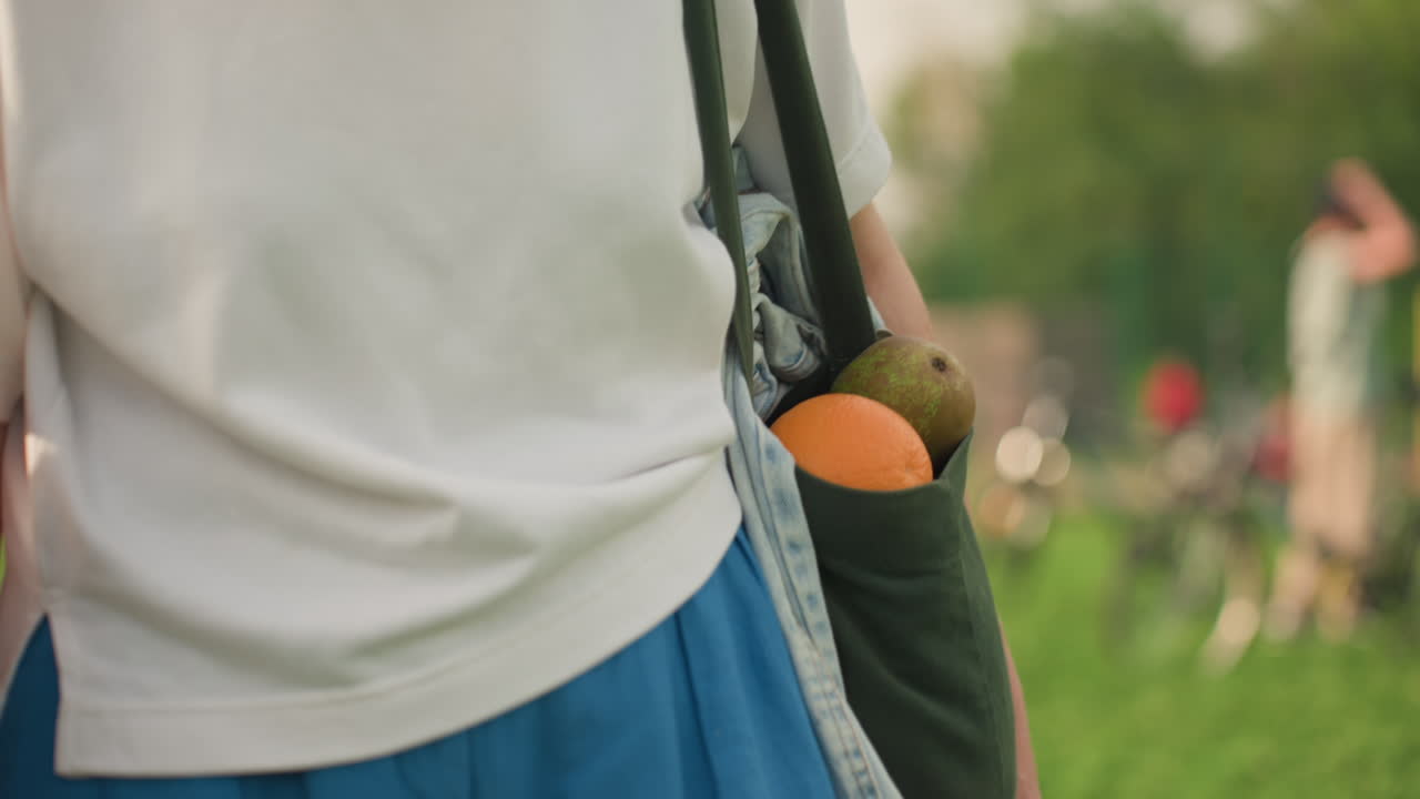 Hand view of picnic mom carrying fruits in black canvas bag while walking through grassy park, denim jacket visible, warm daylight highlighting orange and pear, healthy outdoor lifestyle close up