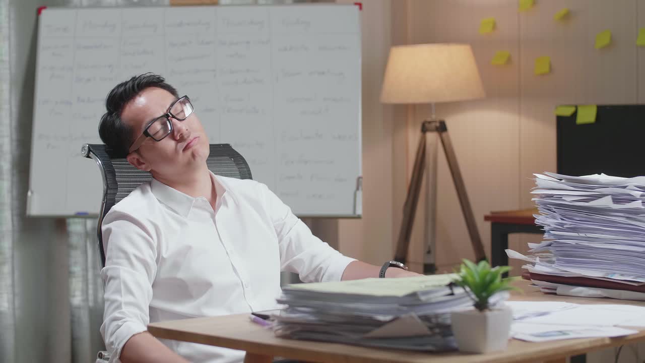 지친 아시아인 남자가 사무실 의자에 기대어 서류로 열심히 일하면서 잠을 자고 있습니다.