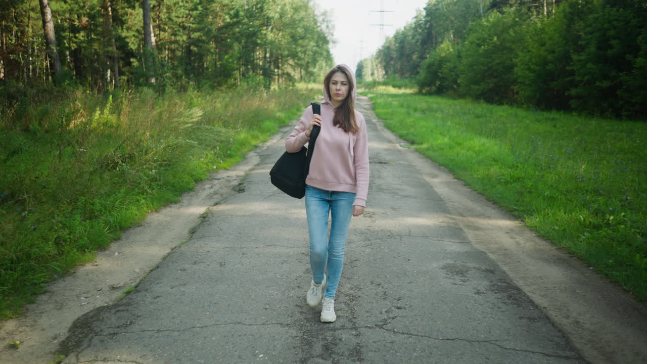 Young lady in pink hoodie walking confidently on cracked rural road, holding black bag strap over shoulder, surrounded by green trees and grass on both sides under clear daylight sky