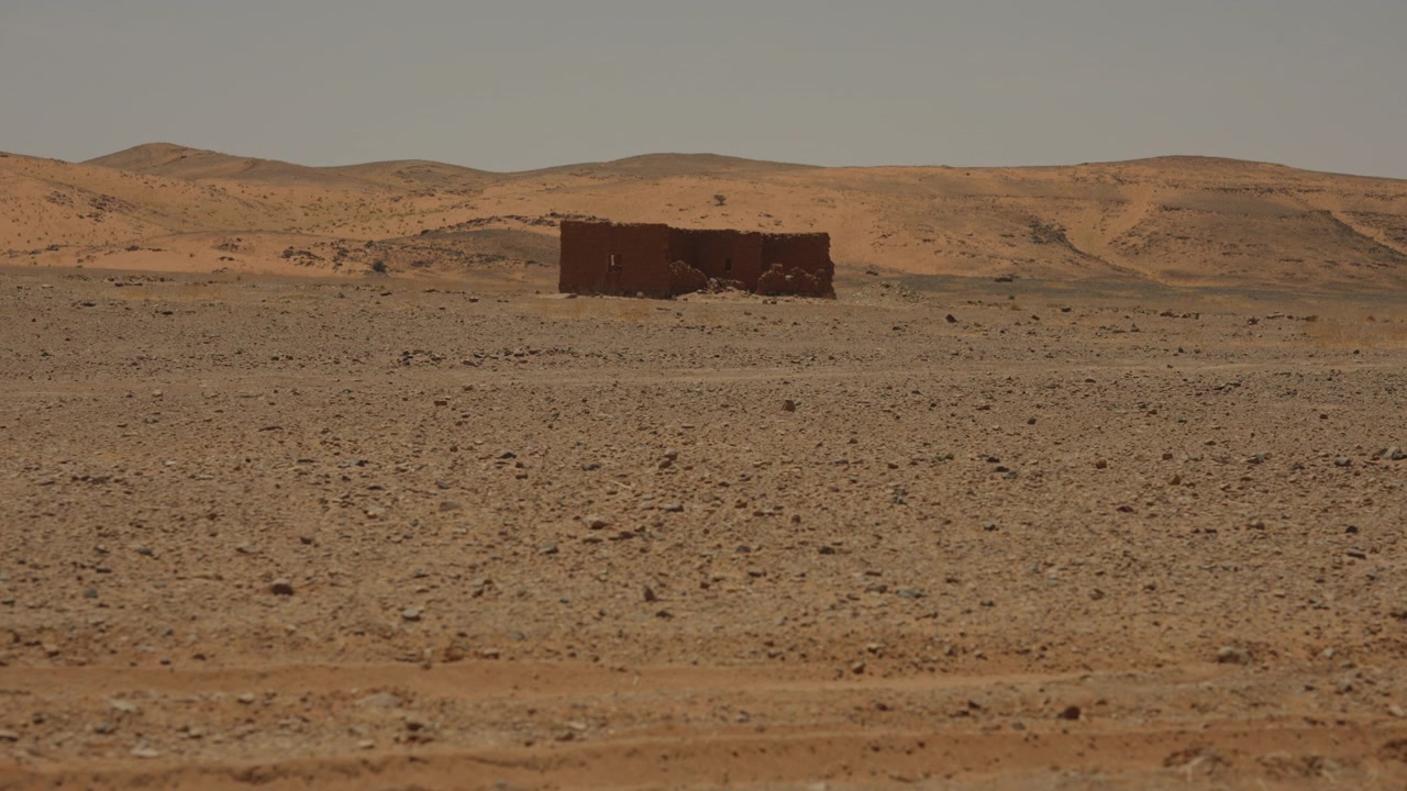 Una singola struttura in mattoni di fango in rovina si erge isolata su una pianura desertica rocciosa con dolci colline di sabbia sullo sfondo.