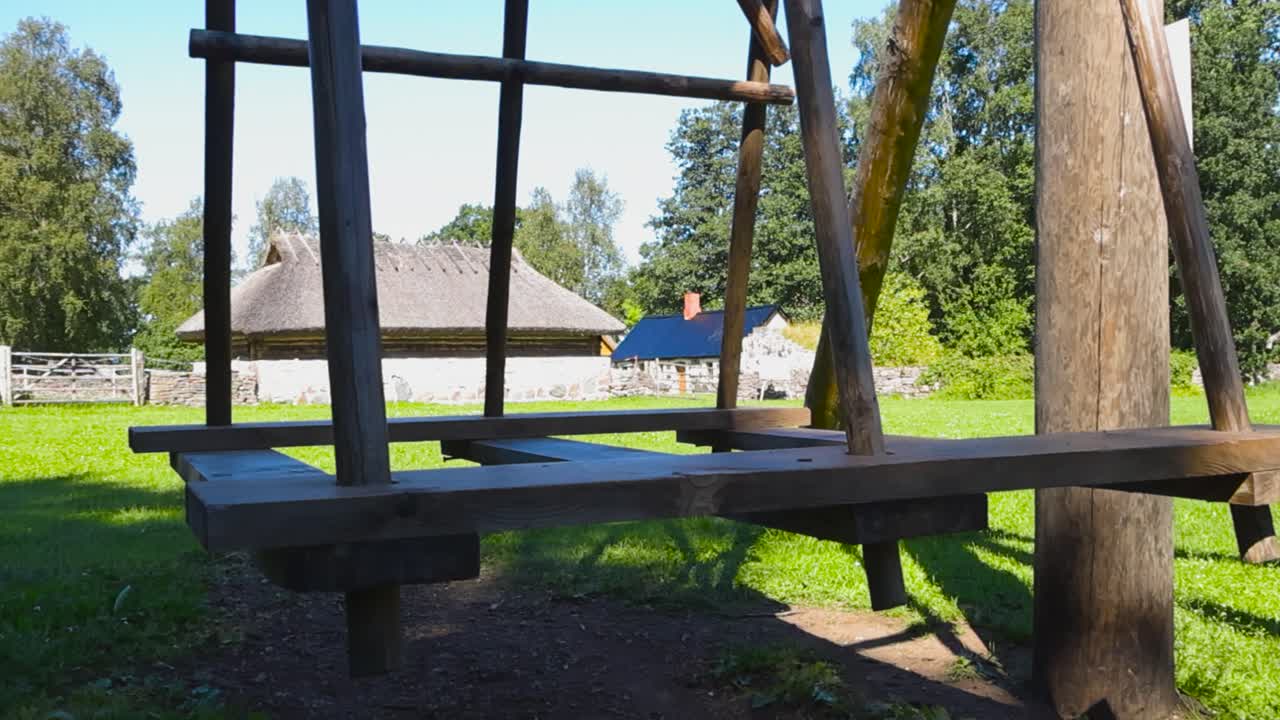 Old traditional and historic wooden village swing in a sunny summer old garden with green grass and old log buildings with thatched straw roof in the bckground. Blue sky and white clouds visible