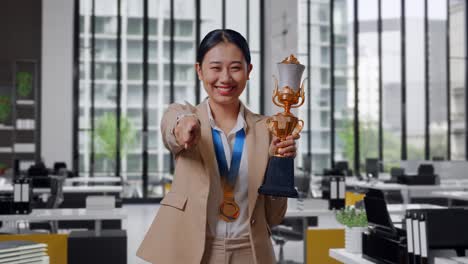 mujer de negocios asiática en traje con una medalla de oro y trofeo tocando su pecho luego sonriendo y señalando a la cámara estando orgullosa de sí misma en la oficina