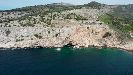 Vista-Aérea-De-Un-Acantilado-Junto-Al-Mar-Con-Agua-Turquesa-Y-Una-Pequeña-Cueva,-Thassos,-Grecia,-Europa