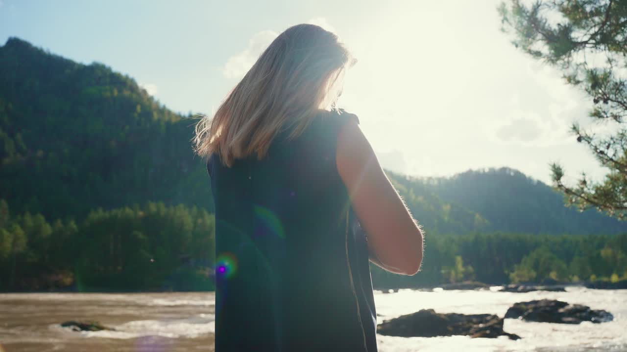 Una donna sta in piedi vicino al fiume, di spalle alla macchina fotografica, osservando l'acqua che scorre. La luce del sole risplende dietro di lei, proiettando una luce soffusa. Indossa un vestito scuro senza maniche, i capelli le scivolano dolcemente. La natura la circonda: alberi, montagne.