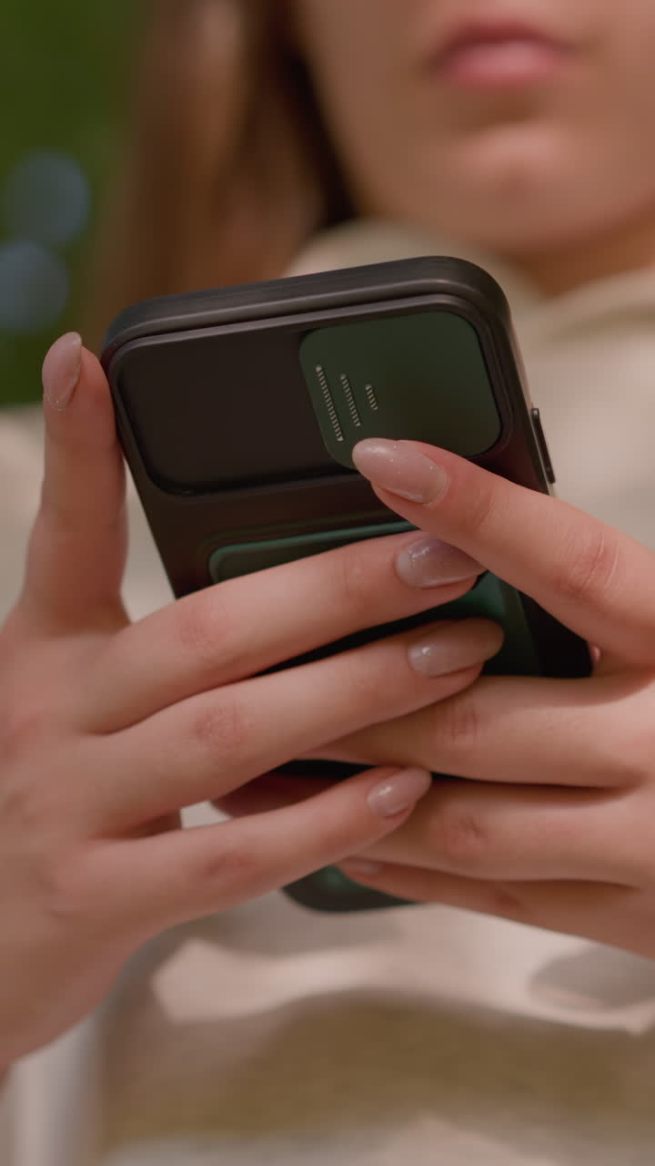 primer plano de enfoque individual operando un teléfono inteligente con uñas pulidas, escribiendo con precisión mientras las sombras caen suavemente sobre la ropa de color claro, con un fondo verde naturalmente borroso