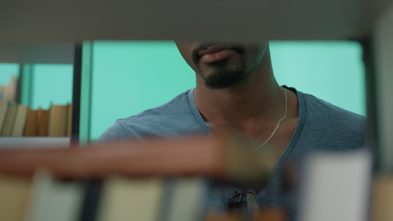 Close up of person wearing casual shirt reaching for book from shelf, partially obscured by rows of books, with turquoise wall in background creating a vibrant indoor library setting