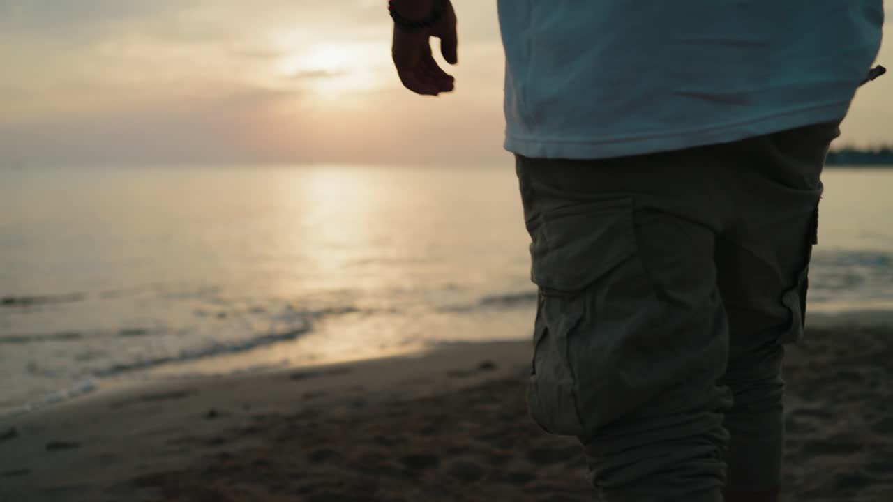Inquadratura media di un uomo che cammina sulla spiaggia verso l'acqua all'alba, ripreso da dietro al rallentatore