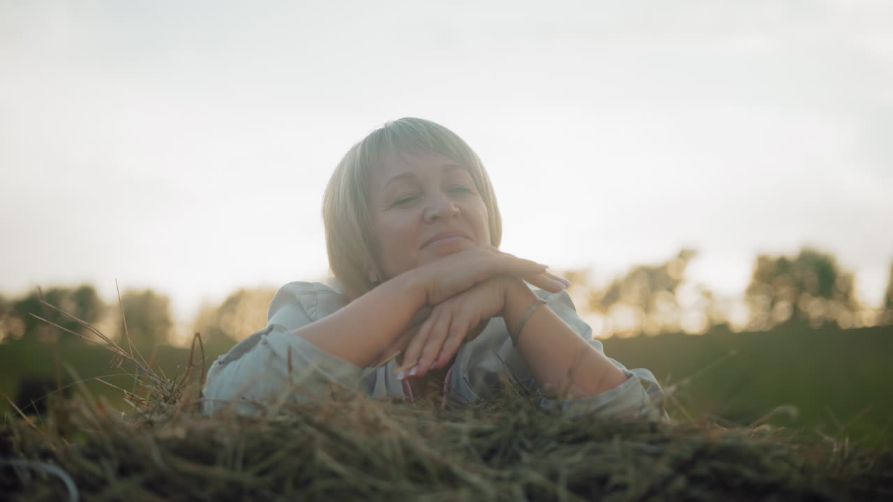 женщина среднего возраста лежит на куче сена с теплой отражающей улыбкой, позирует в открытом поле, мягкий золотой солнечный свет создает мирную сельскую атмосферу с размытым фоном