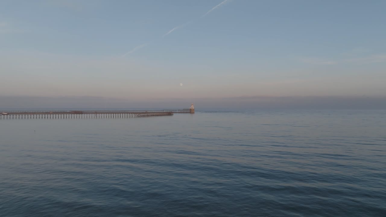 Captivating drone footage of a tranquil sunset over Blyth Lighthouse, with dolphins playing in the calm waters, illuminated under a pastel sky and rising full moon.