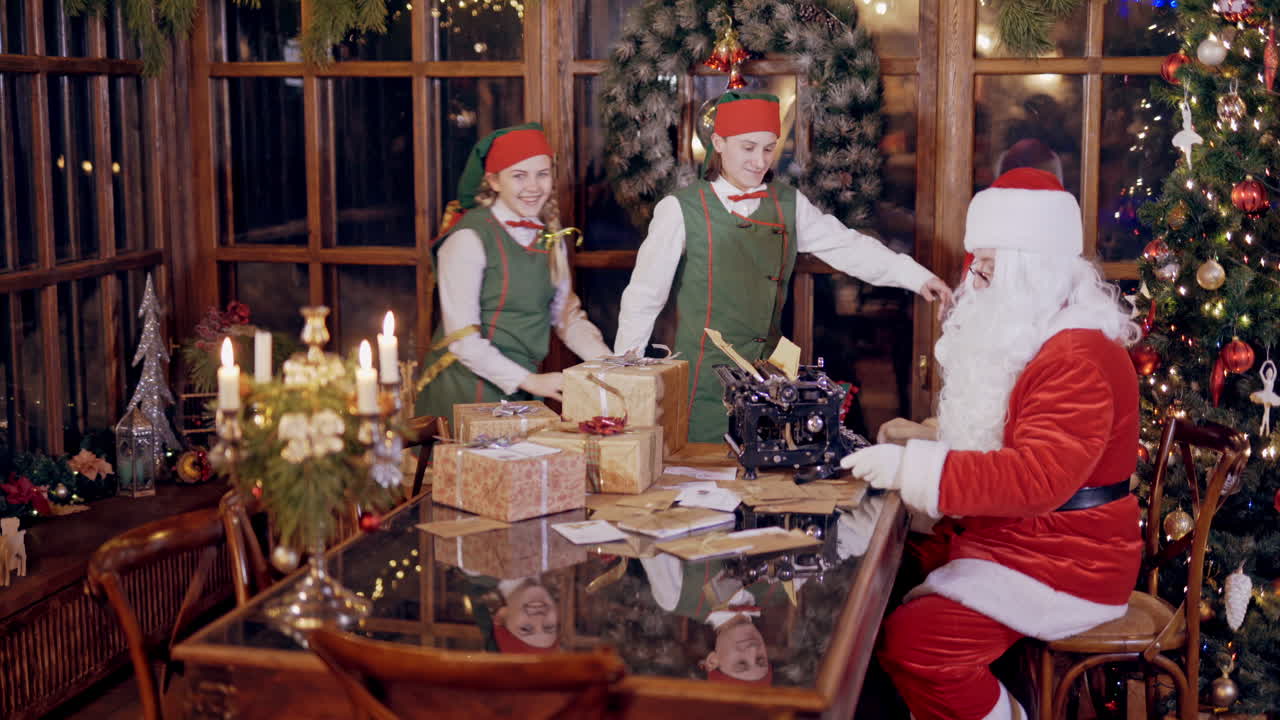 Merry atmosphere with elves and Santa Claus. Santa with white beard in red costume is typing letter on the background of funny elves. New Year's decorations in Santa's residence.